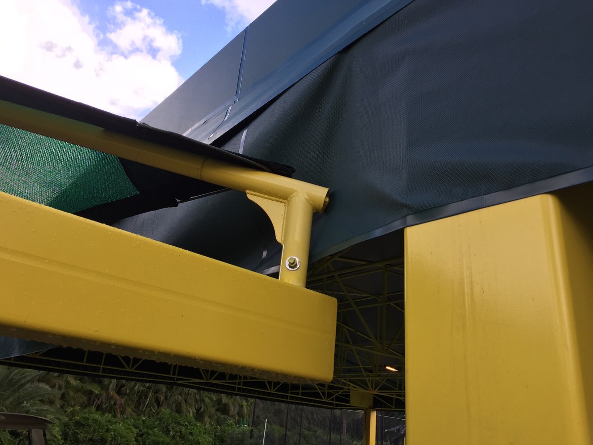 Bolted connection between yellow steel cross-beam and the dark fabric canopy of the park pavilion under a partly cloudy sky.