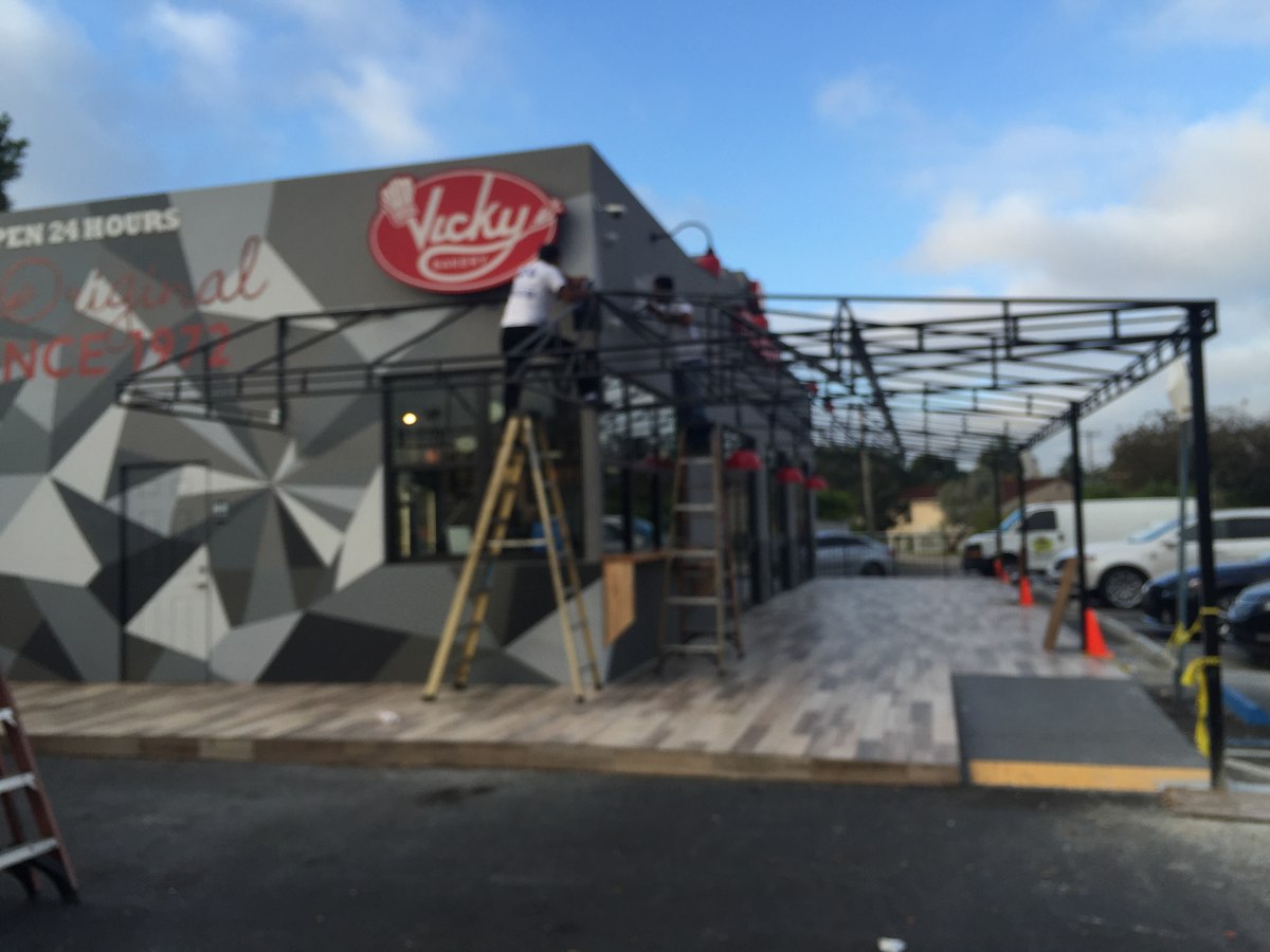 Two crew members on ladders weld up a steel pergola frame over the freshly-paved patio at Vicky Bakery, geometric mural visible.