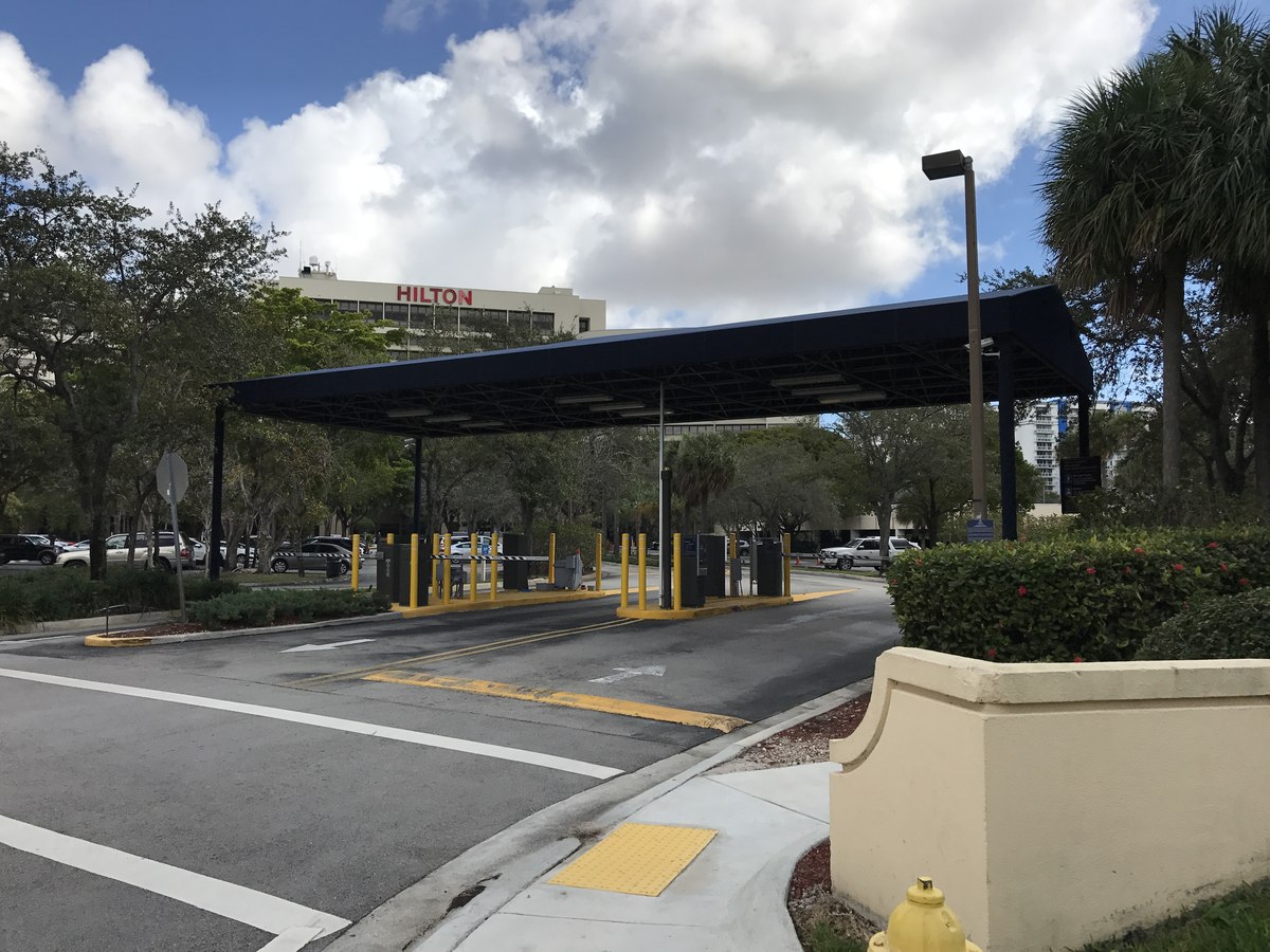 Long navy structural canopy on steel posts spans the entry drive of a Hilton hotel, with the red HILTON sign visible above the trees.