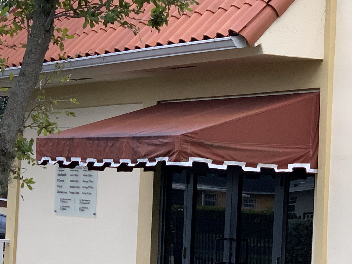 Burgundy commercial-style awning with white scalloped piped trim sheltering an entrance with a posted business-hours sign on a tan stucco facade.