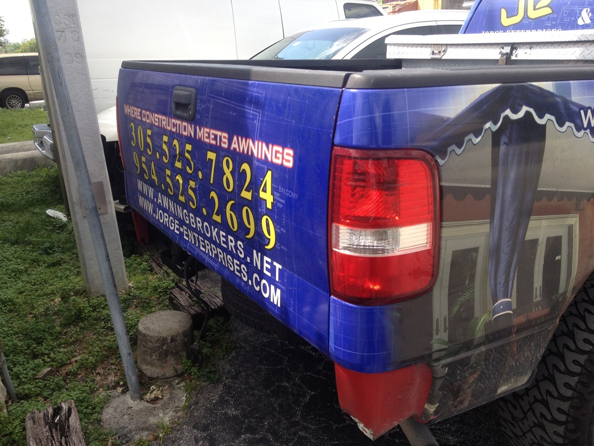 Tailgate of the JE/Awning Brokers blue branded truck reading WHERE CONSTRUCTION MEETS AWNINGS with phone numbers and websites.