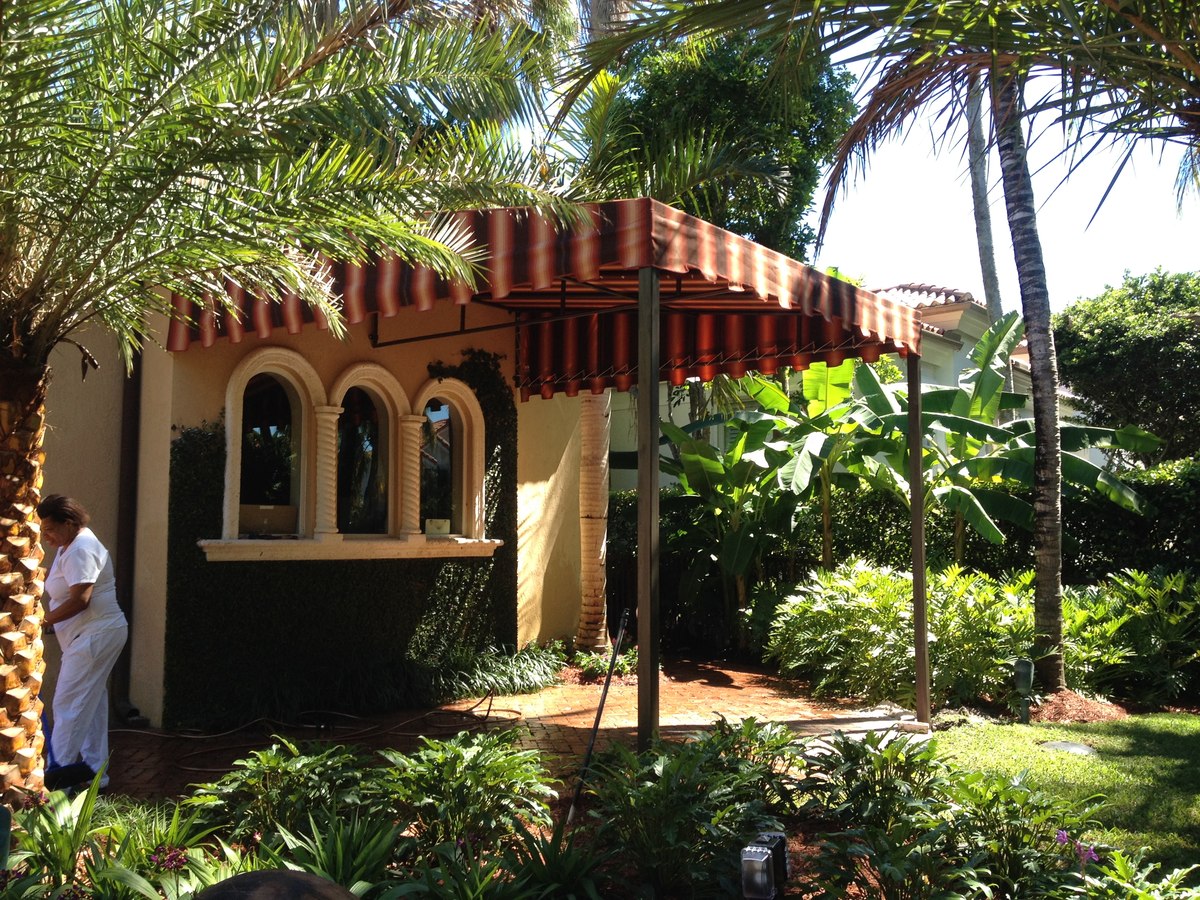 Red-and-tan striped fabric awning over a Mediterranean-style home patio framed by lush palms and tropical landscaping.