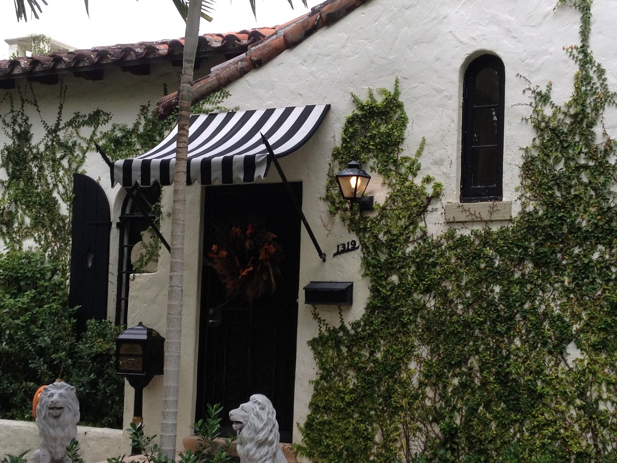 Black-and-white striped fabric awning above a Spanish-tile-roof entry door of an ivy-covered Mediterranean cottage.