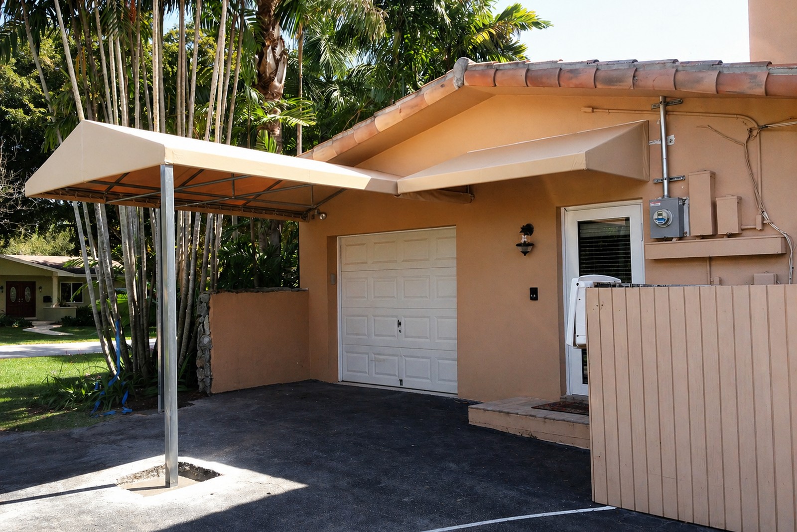 Clean aluminum-frame carport awning extends over a driveway at a SoFla home, palms behind, terra-cotta tile roof beyond.