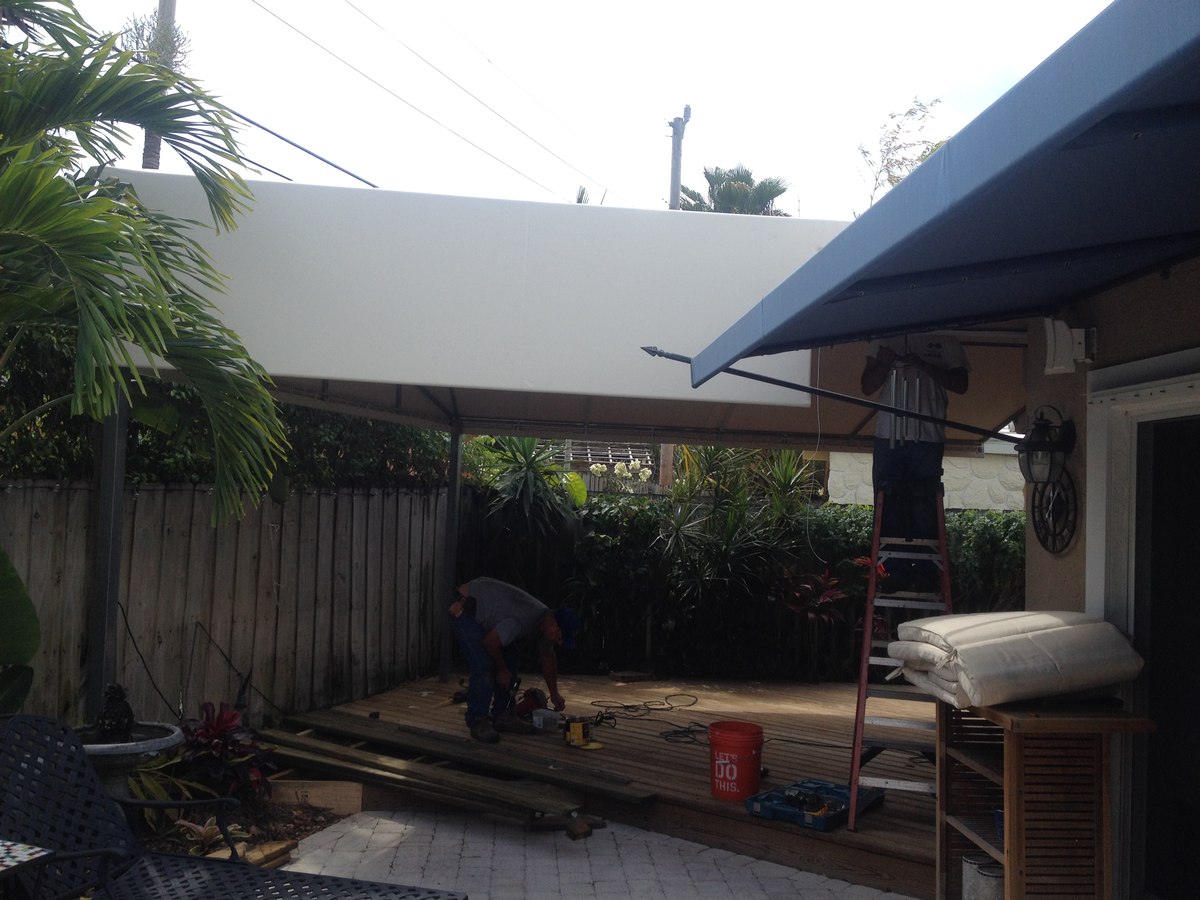 Worker installs a flat white aluminum patio cover next to an existing navy retractable awning in a tropical Florida backyard with deck.