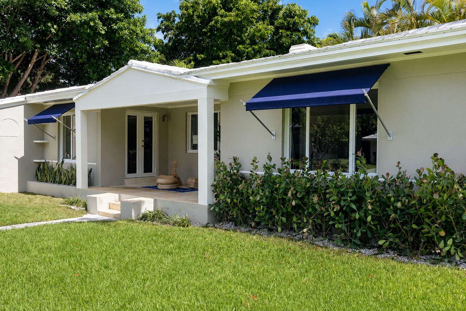 Two matching navy bahama window awnings shade the front of a clean white SoFla ranch home, hedges and palms in front.