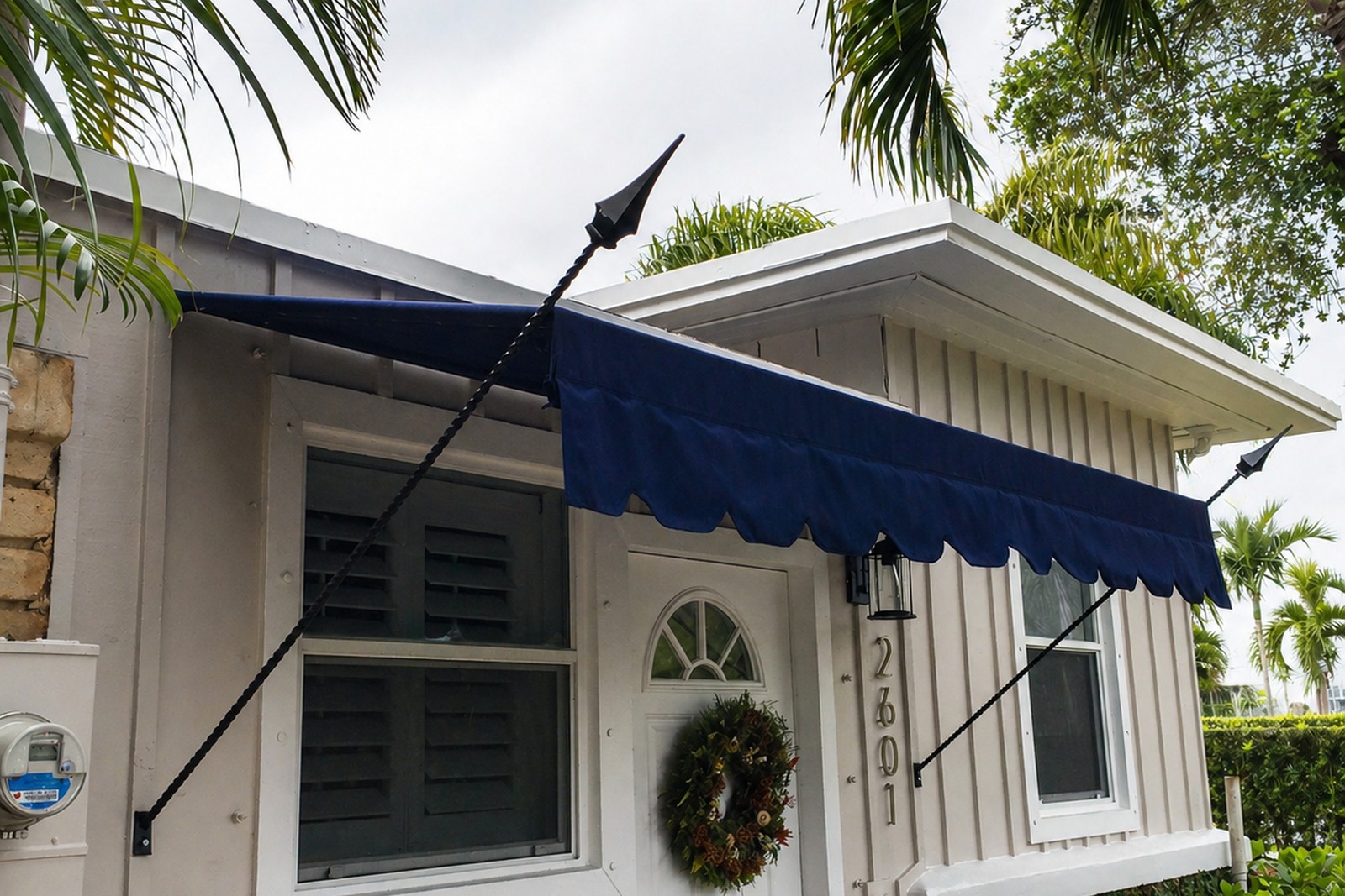Navy scalloped spear-point awning over the front door of a small white board-and-batten cottage, tropical palms behind.
