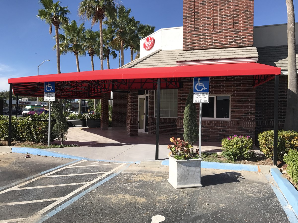 Long red commercial fabric canopy over a brick restaurant entrance, with palm trees and sky overhead.