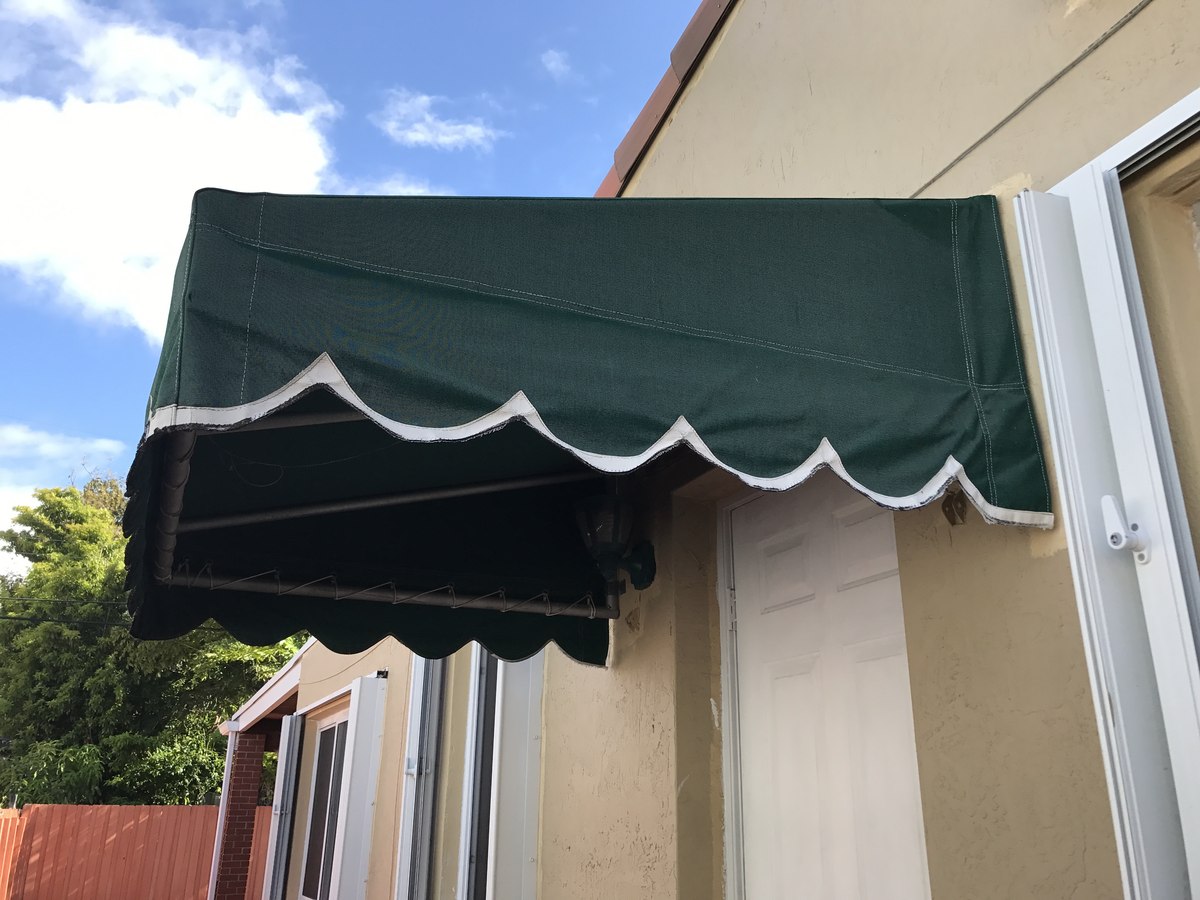 Forest-green awning with white scalloped trim shelters a side door of a tan stucco home, with a fence and tree visible at left.
