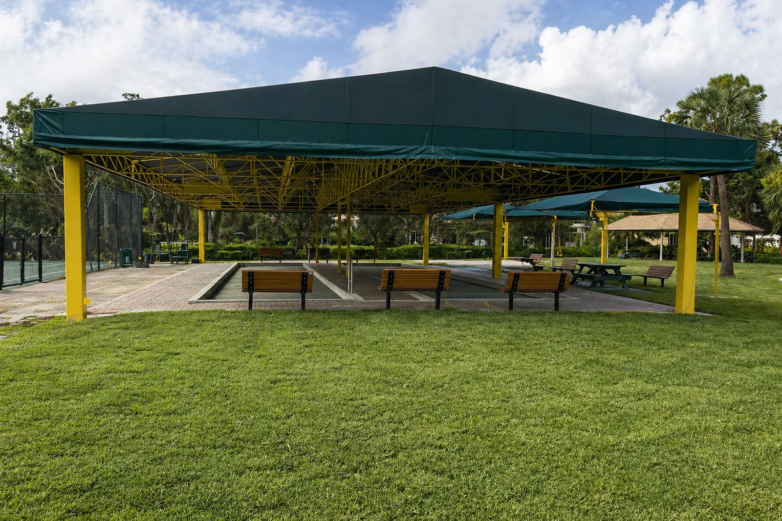 Large commercial shade canopy with dark-green fabric roof on yellow steel posts over benches and bocce courts in a park.