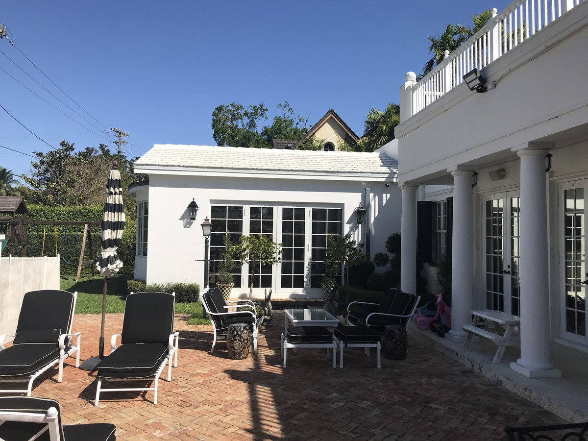 Brick-paver back patio of an upscale white classic home with black-stripe market umbrella, modern lounge furniture, French doors and pool toys.