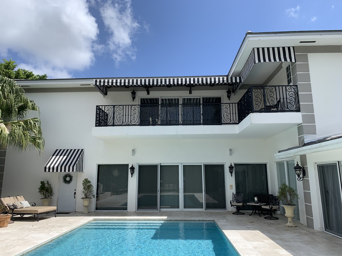 White-stuccoed Mediterranean two-story home with matched black-and-white striped awnings on the upper floor and over a French door, pool in foreground.