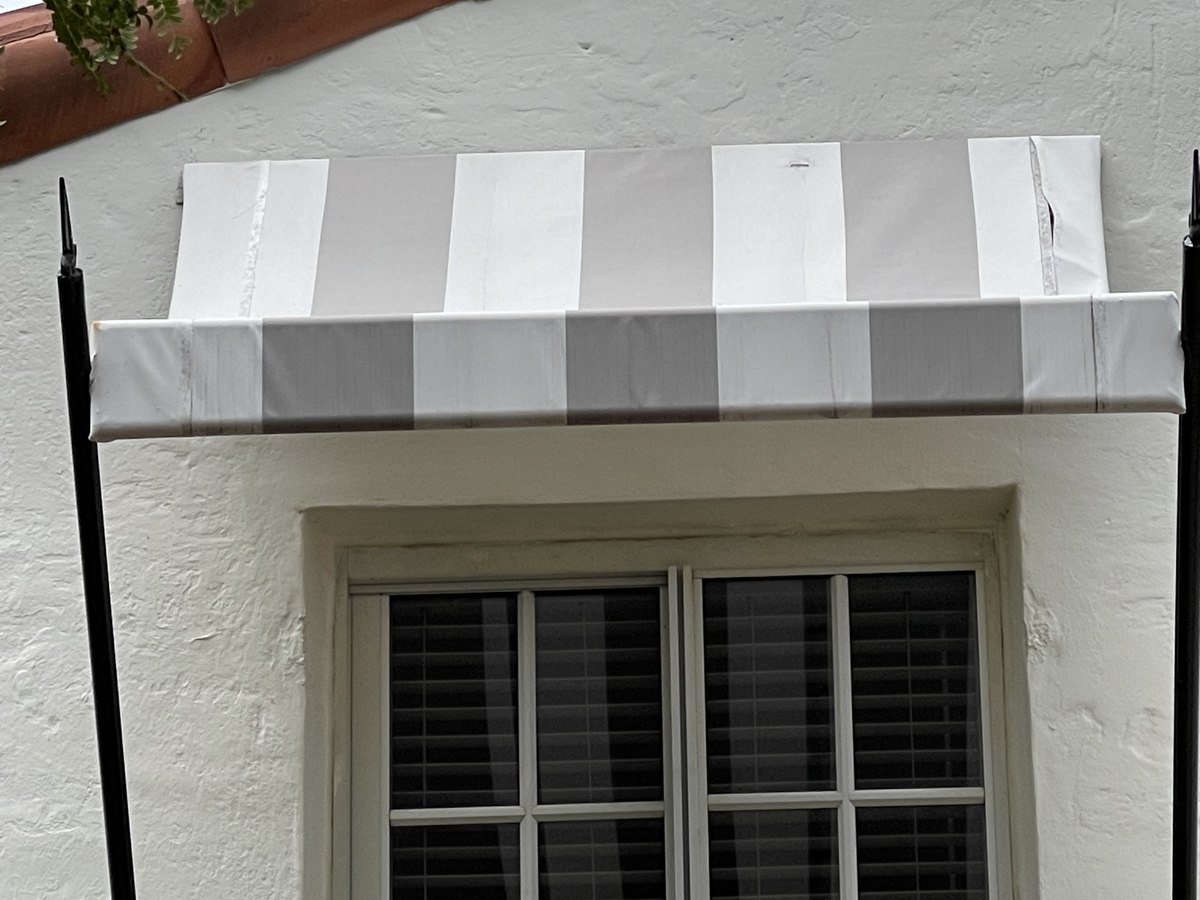 Top-down detail of the grey-and-white striped window awning with iron finials and terra-cotta roof tile in the corner.