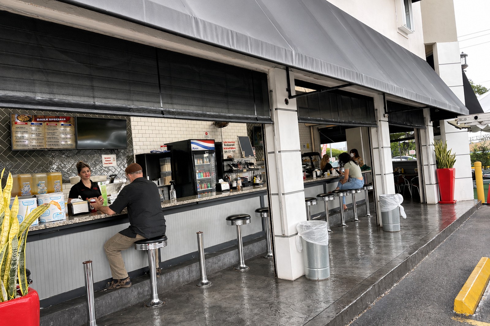 Diner counter open to the sidewalk under a charcoal-grey commercial awning; customers on chrome stools order at the window.