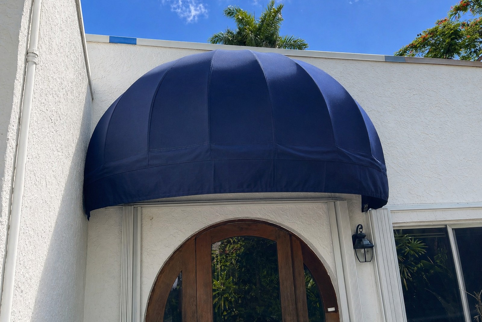 Solid navy dome awning over an arched wood front door at a stucco SoFla home, palms framing the entry.