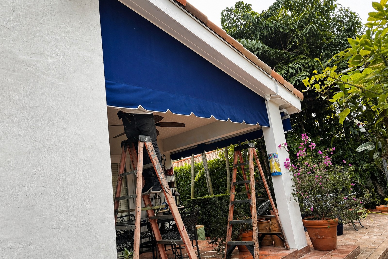 Crew member on a wooden ladder installs a navy fixed dome awning over a tropical patio with bistro furniture and bougainvillea in bloom.