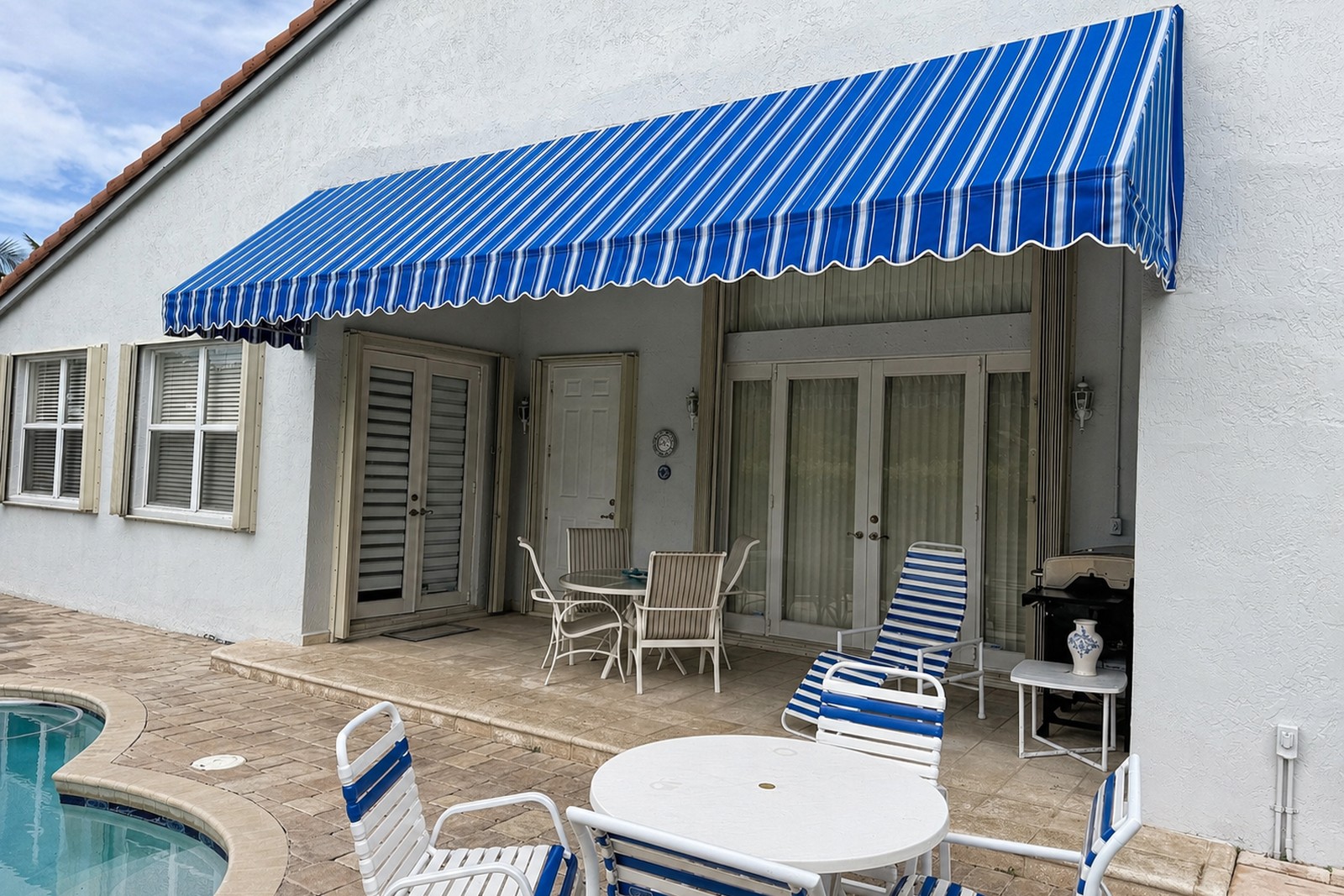 Bold blue-and-white striped retractable awning shades a SoFla pool patio with strap chairs, French doors, and louvered shutters.