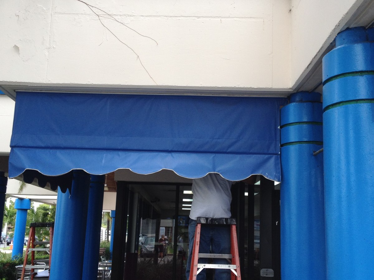 Worker on a ladder installing or maintaining a navy-blue commercial canopy with scalloped valance over a storefront entry framed by blue painted columns.