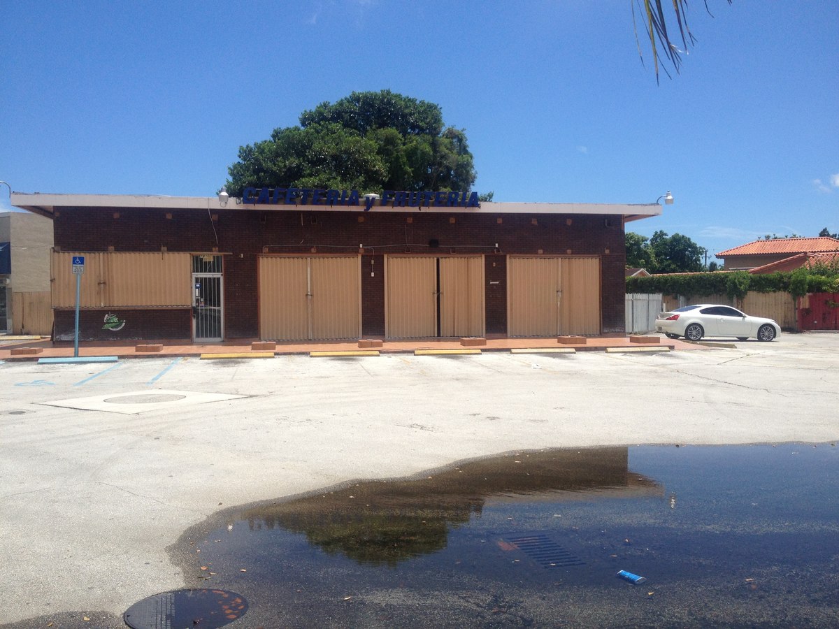 Cafeteria y Fruteria storefront with full-width tan accordion hurricane shutters closed across the facade, parking lot in front.