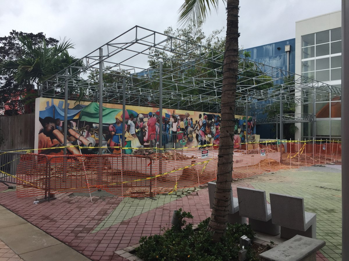 Steel canopy frame staged over a colorful Caribbean-themed mural at the Haitian Cultural Center, pavers torn up beneath.