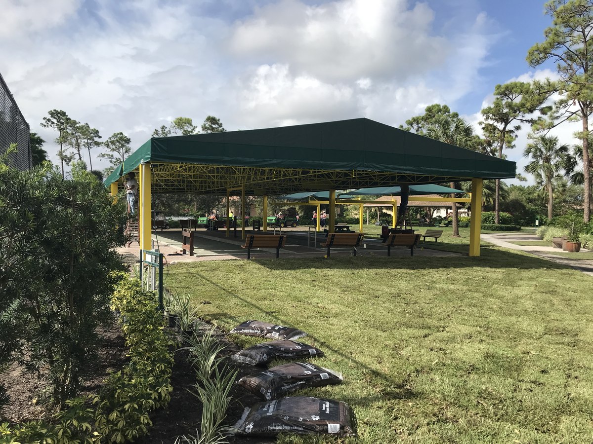 Wide finished view of the yellow-truss green-canopy park pavilion over bocce courts framed by tropical landscaping under a partly cloudy sky.