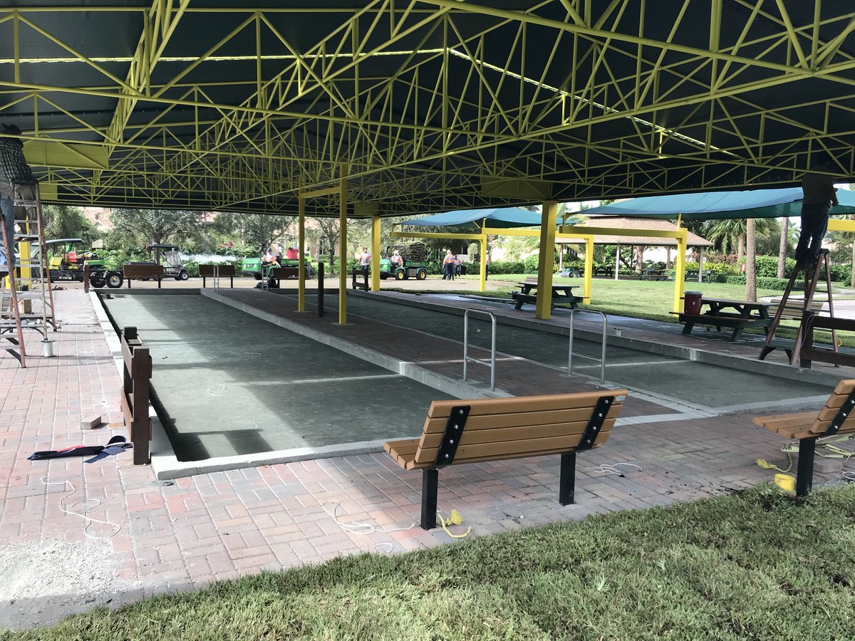 View from inside the pavilion looking down a row of bocce courts and benches under the yellow-truss canopy.