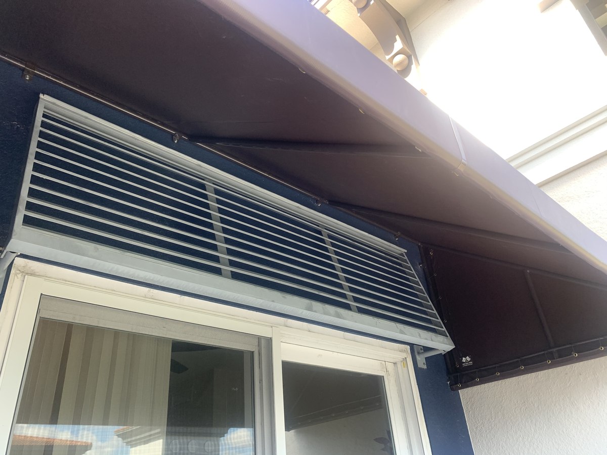 Navy-blue Bahama-style louvered hurricane shutter mounted at an angle over a residential window, with matching navy side wall.